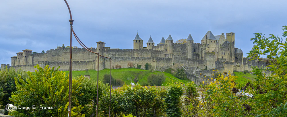 Excursion from Toulouse to Carcassonne: view of the fortified walls of the medieval city, surrounded by greenery under a cloudy sky. Photo by diegoenfrance.com
