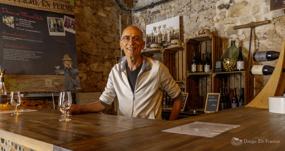 Portrait du vigneron Marco Paulo derrière le comptoir de sa cave à vin traditionnelle, entouré de bouteilles, d'histoire familiale et d'objets anciens. Photo par diegoenfrance.com