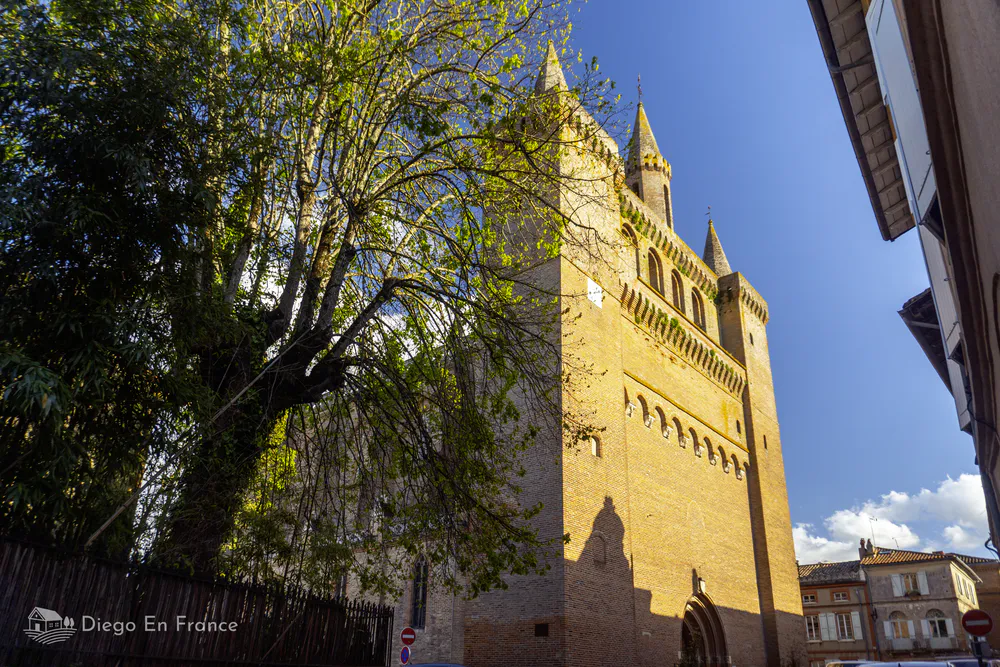 Gothic brick church in Saint-Sulpice-la-Pointe seen from the street, next to a leafy tree.