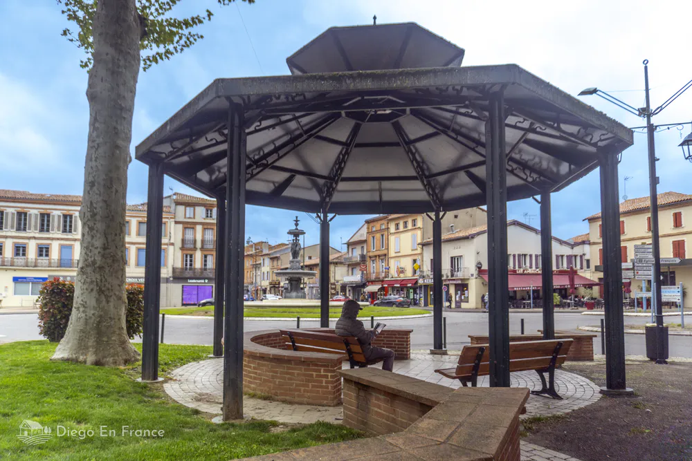A gazebo in the central square of Lavaur, surrounded by pink brick houses and benches under the shade of trees.
