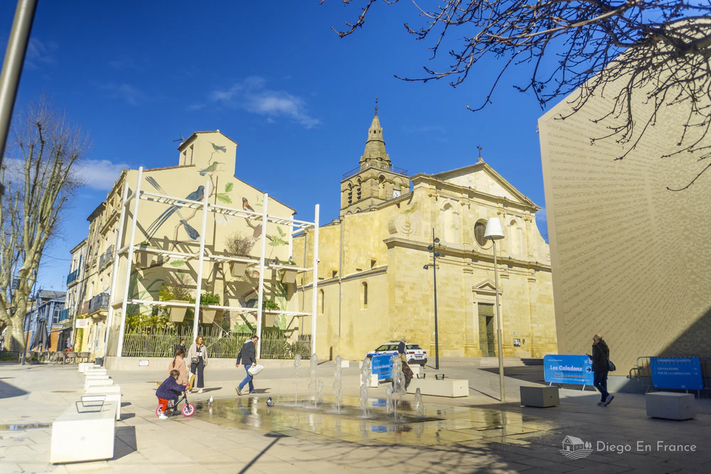 Vue de la place Louis Rey à Lunel avec sa fresque artistique, sa fontaine pour enfants et l'église principale de la ville. Photo par diegoenfrance.com