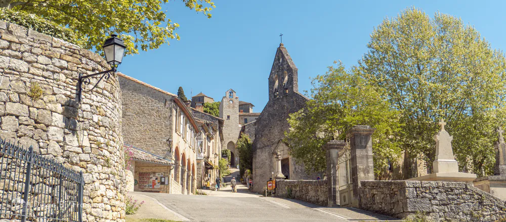 View of the church and bell tower of Bruniquel framed by trees, stone walls, and village life.