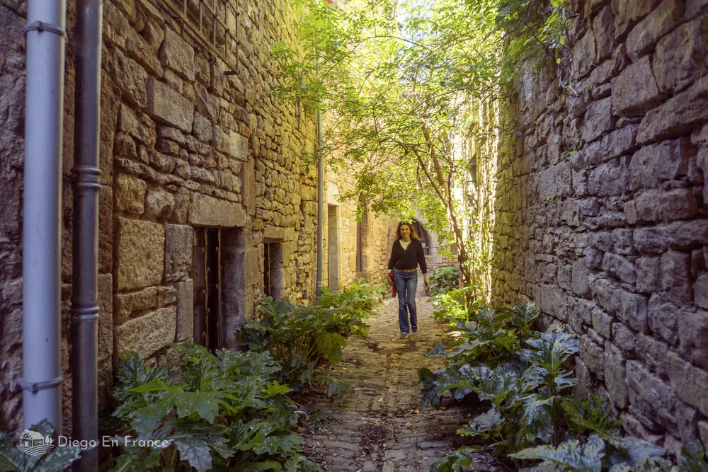 Woman walking through a narrow, leafy medieval passageway lined with stone walls in Bruniquel.